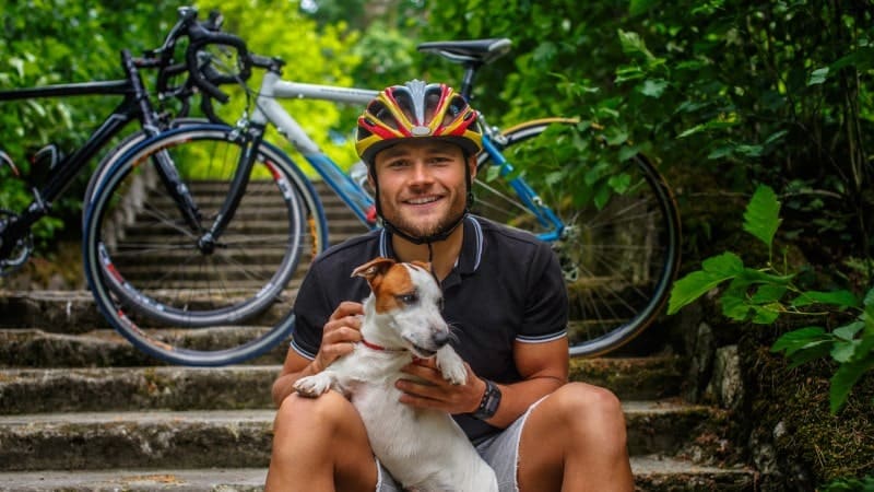 A man wearing a cycling helmet and black cycling attire sits on outdoor steps, smiling and holding a small white and brown dog. Behind him are two bicycles resting against the steps.