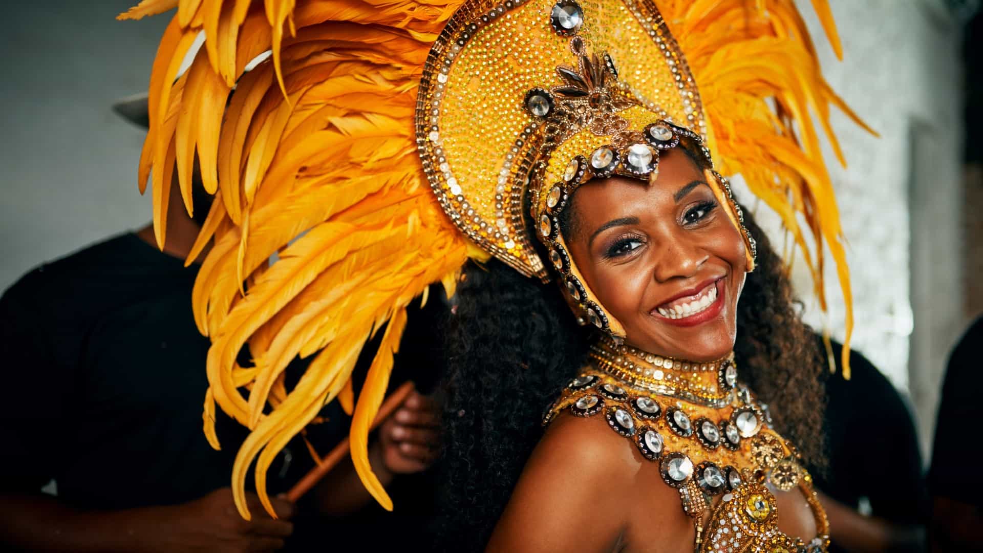A woman dressed in a vibrant carnival costume smiles brightly. Her outfit features an elaborate headdress adorned with large yellow feathers and sparkling gems, complementing her ornate golden attire decorated with sequins and stones. The background is blurred, focusing attention on her joyful expression and festive attire.