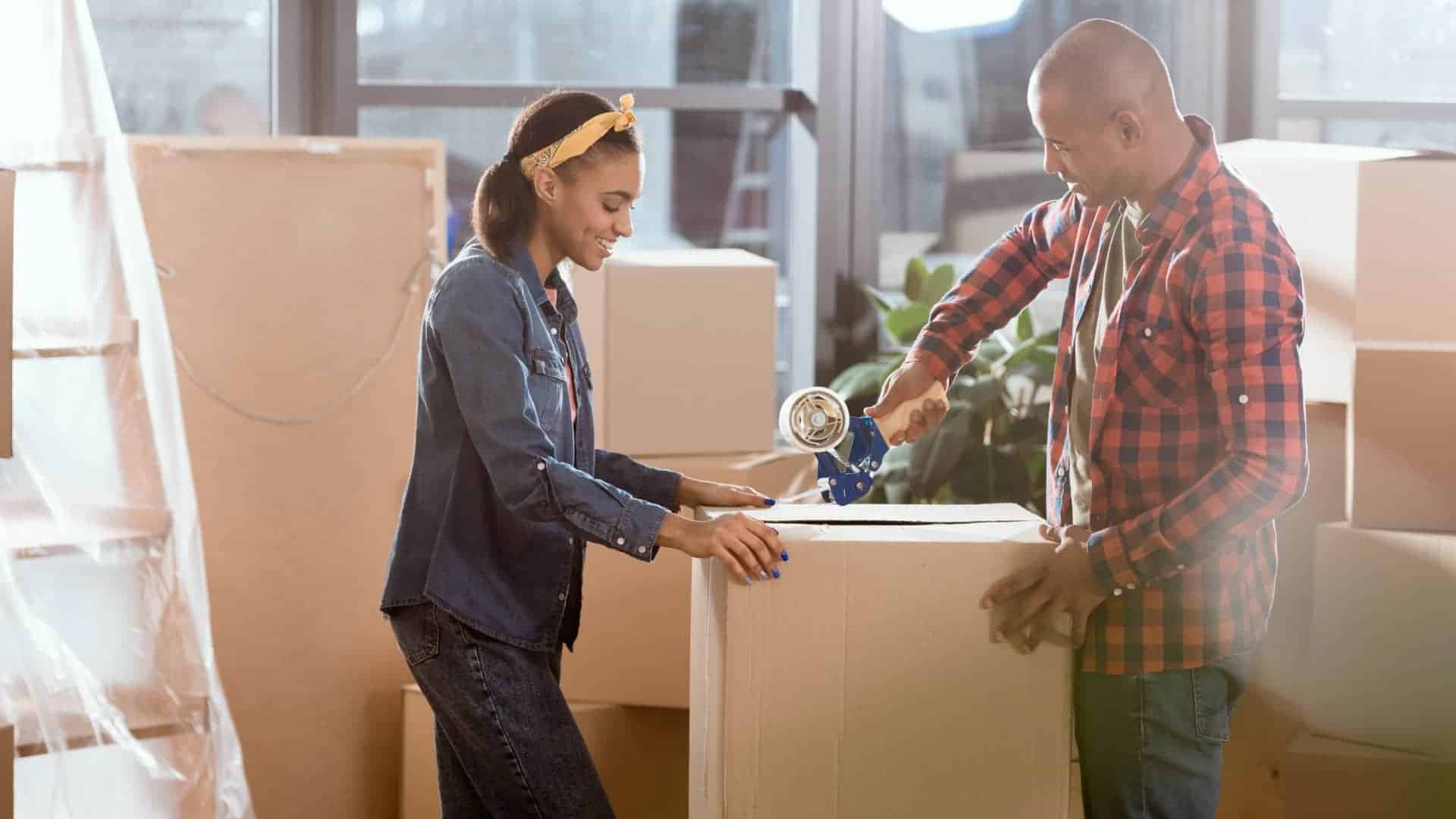 A couple packing boxes together in a brightly lit room. The woman, wearing a denim shirt and yellow headband, holds the box steady while the man, dressed in a red and black plaid shirt, seals it with packing tape. The room is filled with cardboard boxes, indicating a move or relocation.