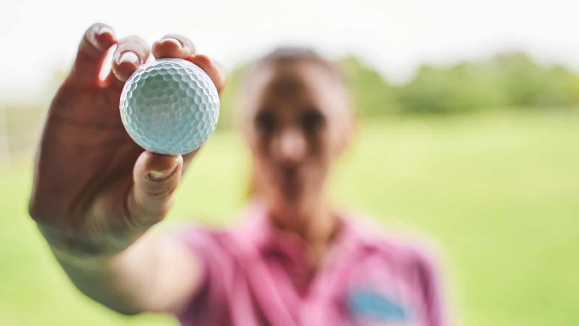 The image shows a woman holding a golf ball close to the camera, with the ball in sharp focus and the woman blurred in the background. She appears to be wearing a pink shirt and is outdoors, likely on a golf course. The focus on the ball highlights its texture and dimples.