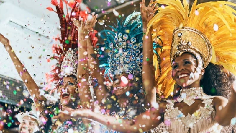 A group of women celebrate at a carnival, all dressed in extravagant costumes. Each woman wears a unique headdress adorned with feathers and jewels—one in yellow, another in blue, and another in red. They are surrounded by a shower of confetti, their arms raised in joy and excitement. The festive atmosphere is vibrant and filled with energy, capturing the essence of a lively carnival celebration.