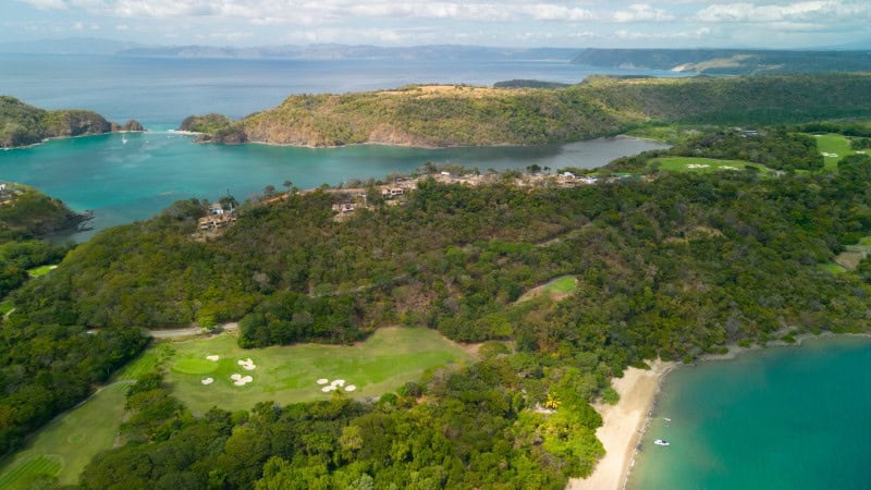 The image is an aerial view of a scenic coastal area featuring a golf course surrounded by lush greenery. The coastline includes pristine beaches with turquoise waters, and there are hilly terrains covered with dense trees. The golf course is visible with its green fairways and bunkers, blending beautifully with the natural landscape. In the background, there are more hills and a calm, expansive body of water, likely a bay or ocean, under a partly cloudy sky.