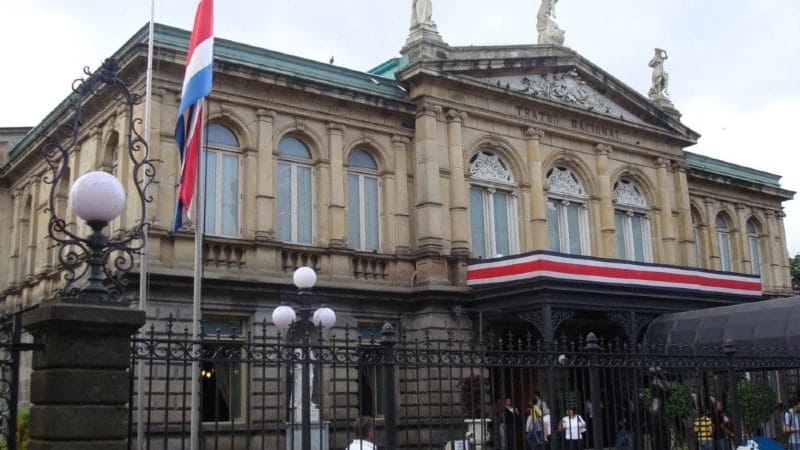 A historic building with ornate architectural details, featuring arched windows and decorative sculptures on the roofline. The façade is adorned with columns and intricate stonework. A large Costa Rican flag is displayed prominently at the front, and a red, white, and blue ribbon drapes across the entrance, signifying national pride. The building is surrounded by a wrought-iron fence with vintage-style lampposts.