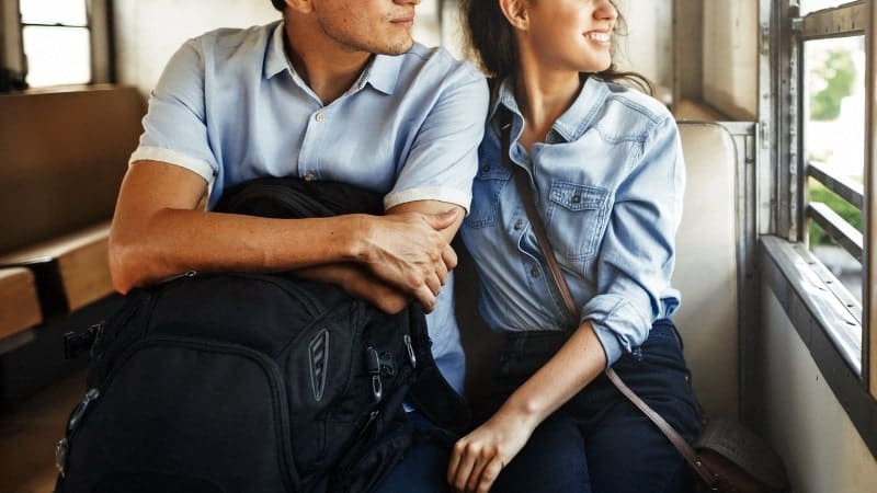 A young couple sits together on a train, both looking out the window with smiles on their faces. They are dressed casually in light blue shirts and dark pants, with the man holding a black backpack on his lap. The sunlight streams through the window, illuminating their faces and creating a warm, relaxed atmosphere. 