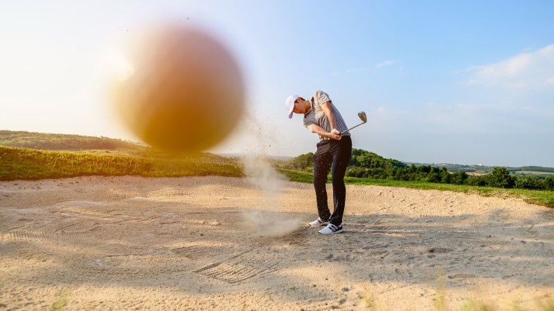 The image captures a golfer in mid-swing, hitting a shot from a sand bunker. The golfer is wearing a white cap, a gray shirt, black pants, and white golf shoes. The focus of the shot is on the golfer's powerful swing, with the sand flying and the golf ball prominently blurred in motion, indicating a strong impact. The background shows a lush green landscape under a clear blue sky, suggesting a sunny day on the golf course.