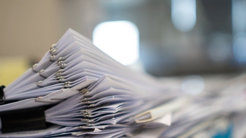 A close-up view of a large stack of paper documents held together with metal binder clips. The focus is on the edges of the papers, creating a blurred background with indistinct shapes and light.