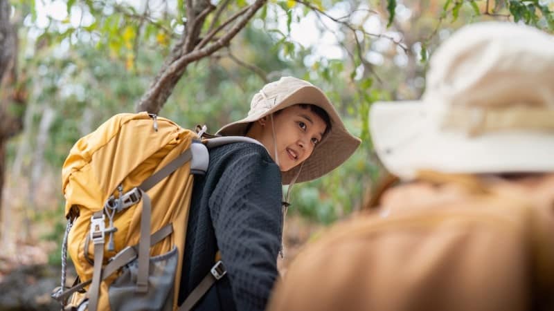 A young hiker wearing a wide-brimmed hat and a black long-sleeve shirt is carrying a large yellow backpack. He looks back with a smile at a fellow hiker, whose back and hat are visible in the foreground.