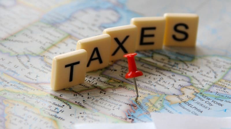 A close-up view of a map with a red pushpin marking a specific location. The word "TAXES" is spelled out in capital letters using Scrabble tiles, arranged neatly on the map.