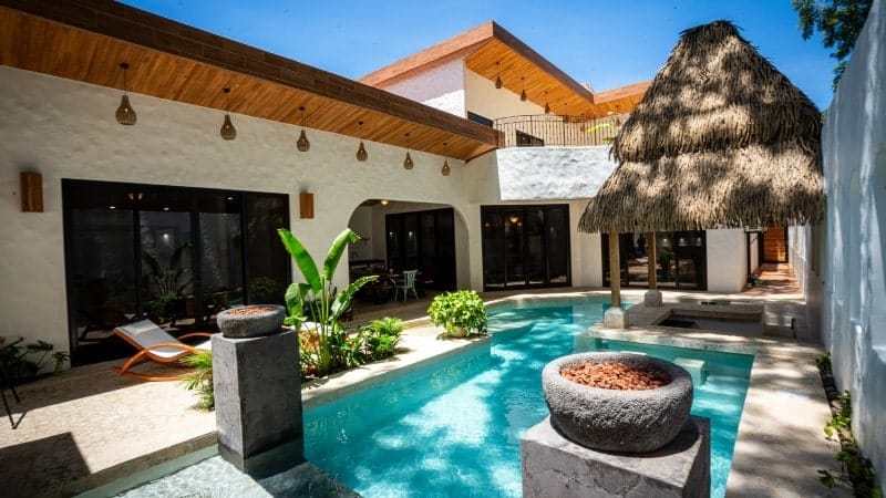 A luxurious outdoor patio area featuring a sparkling blue swimming pool surrounded by lush greenery and tropical plants. The patio is part of a modern house with large glass doors and windows, and the wooden ceiling is adorned with hanging lights. A thatched-roof gazebo stands near the pool, providing a shaded seating area. Comfortable lounge chairs are placed near the pool, offering a relaxing atmosphere. 
