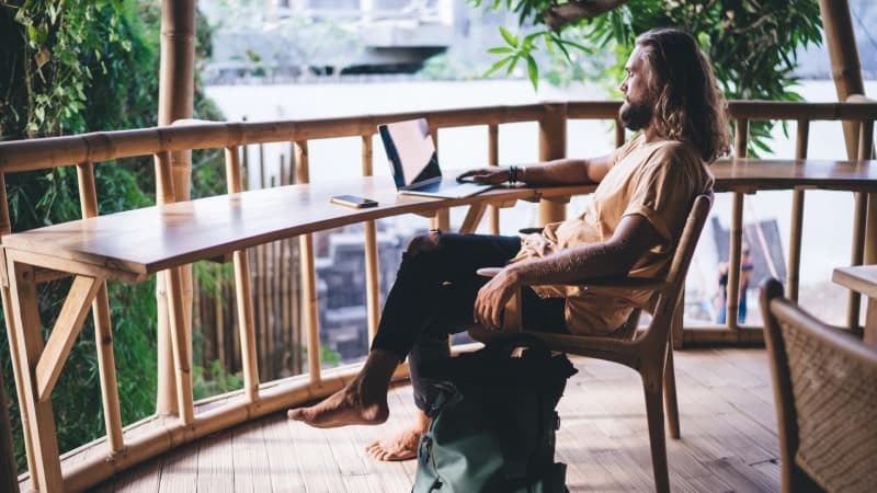 A man with long hair and a beard, wearing a light tan shirt and black jeans, sits barefoot at a bamboo desk on an outdoor balcony. He is working on a laptop, with a smartphone and a notebook placed on the desk. The balcony is surrounded by lush green foliage, providing a serene and natural environment. The desk and chair are made of bamboo, complementing the rustic, eco-friendly setting.