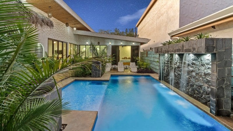 A luxurious outdoor swimming pool area at night, featuring a modern pool with clear blue water and a cascading stone waterfall on one side. The pool is surrounded by lush tropical plants and illuminated by soft lighting. The adjacent house has large glass doors and windows, allowing a view of a well-lit, stylish interior. The wooden ceiling of the patio is adorned with hanging lights, enhancing the serene and inviting ambiance. The night sky is visible above, adding a touch of tranquility to this elegant outdoor space.